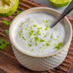 Homemade lime crema in a white bowl with a spoon and squeezed limes.