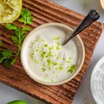 Bowl of lime crema with a spoon in it on a clay platter with lime and cilantro garnish.