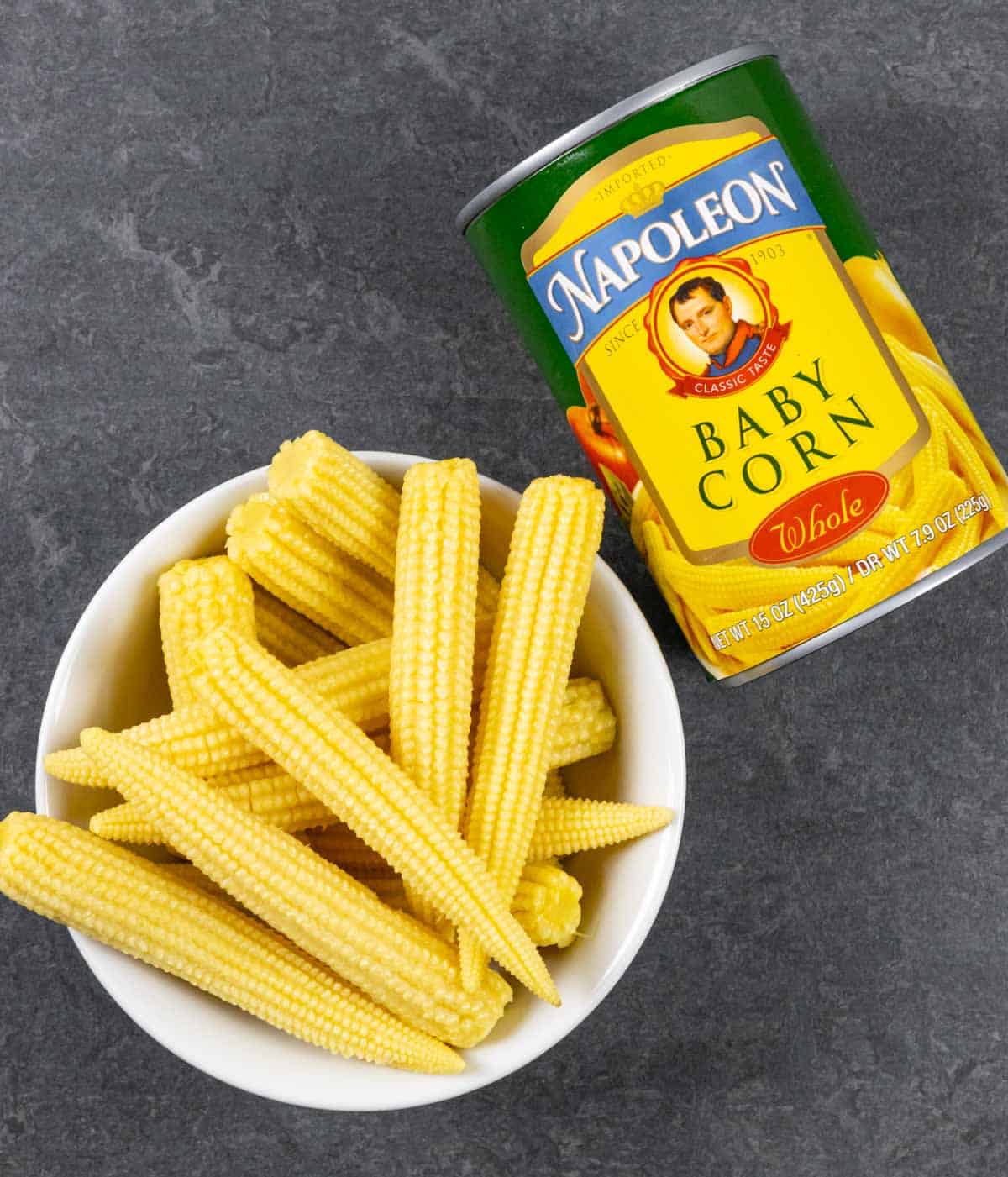 White bowl of baby corns out of the can laying next to it on a grey board.