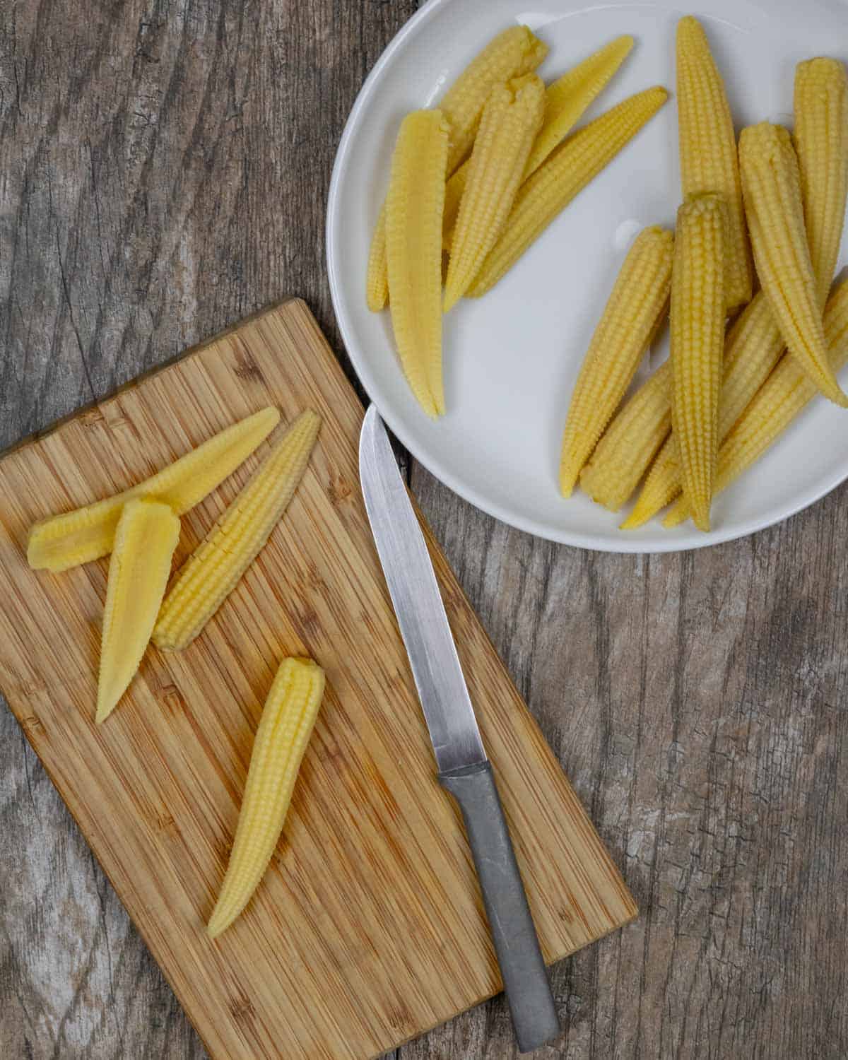 Baby corn on a cutting board sliced in half along it's length with cut and uncut corns on a white plate.