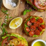 Avocado toast with tomatoes on three different breads on a board with olive oil, lemon wedge and seasonings.