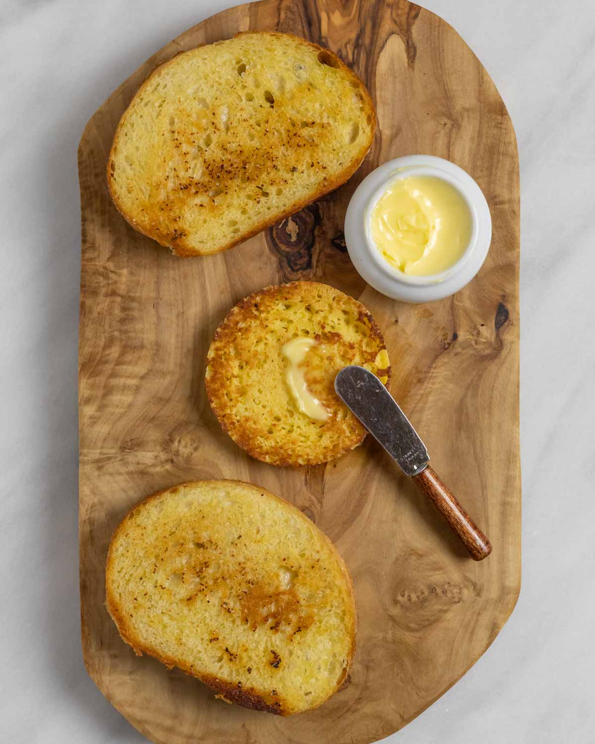 Three slices of toasted bread on a wood board with a ramekin of butter and small spreading knife.
