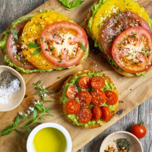 Healthy avocado toast with sliced yellow, red and cherry tomatoes on a board with herbs and spices.