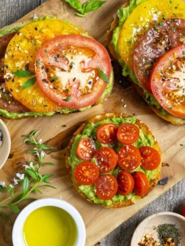 Healthy avocado toast with sliced yellow, red and cherry tomatoes on a board with herbs and spices.