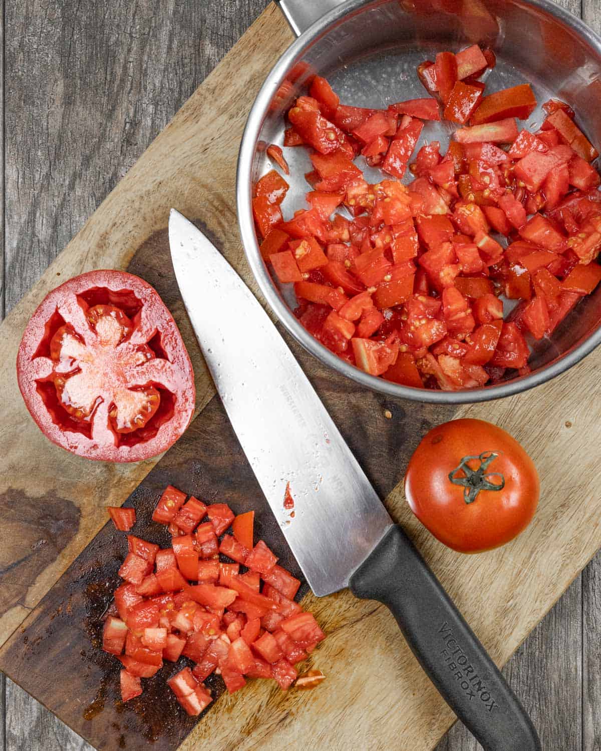 Tomatoes on a board with a chefs knife, some chopped small and in a pan, some on the board and one whole.