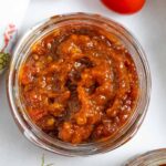 Small glass jar of tomato chilli jam on a board with whole tomatoes next to it.