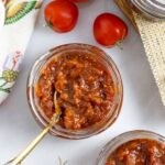 Mason jar of tomato chilli jam with a golden spoon in it, on a board with whole tomatoes.