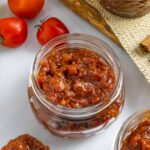 Two clear jars of tomato jam on a marble board with 3 tomatoes.