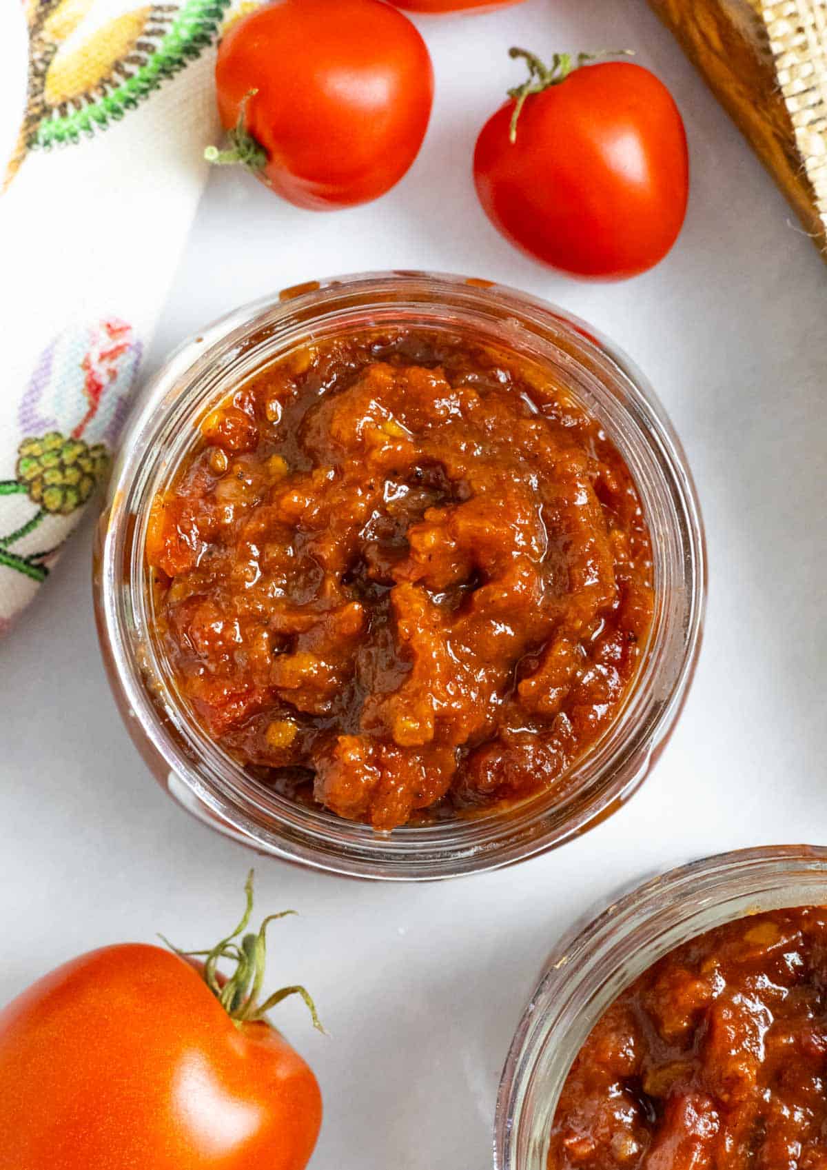 Small glass jar of tomato chilli jam on a board with whole tomatoes next to it.