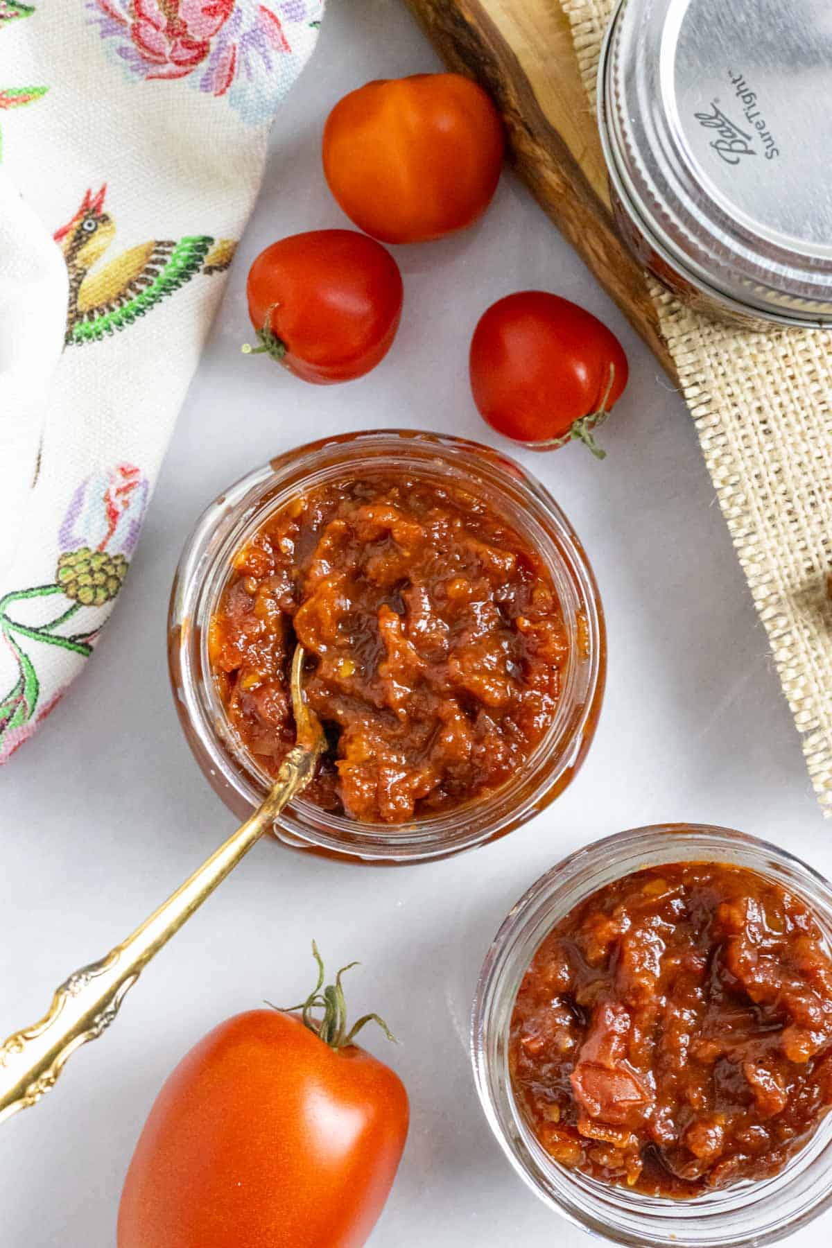 Mason jar of tomato chilli jam with a golden spoon in it, on a board with whole tomatoes.