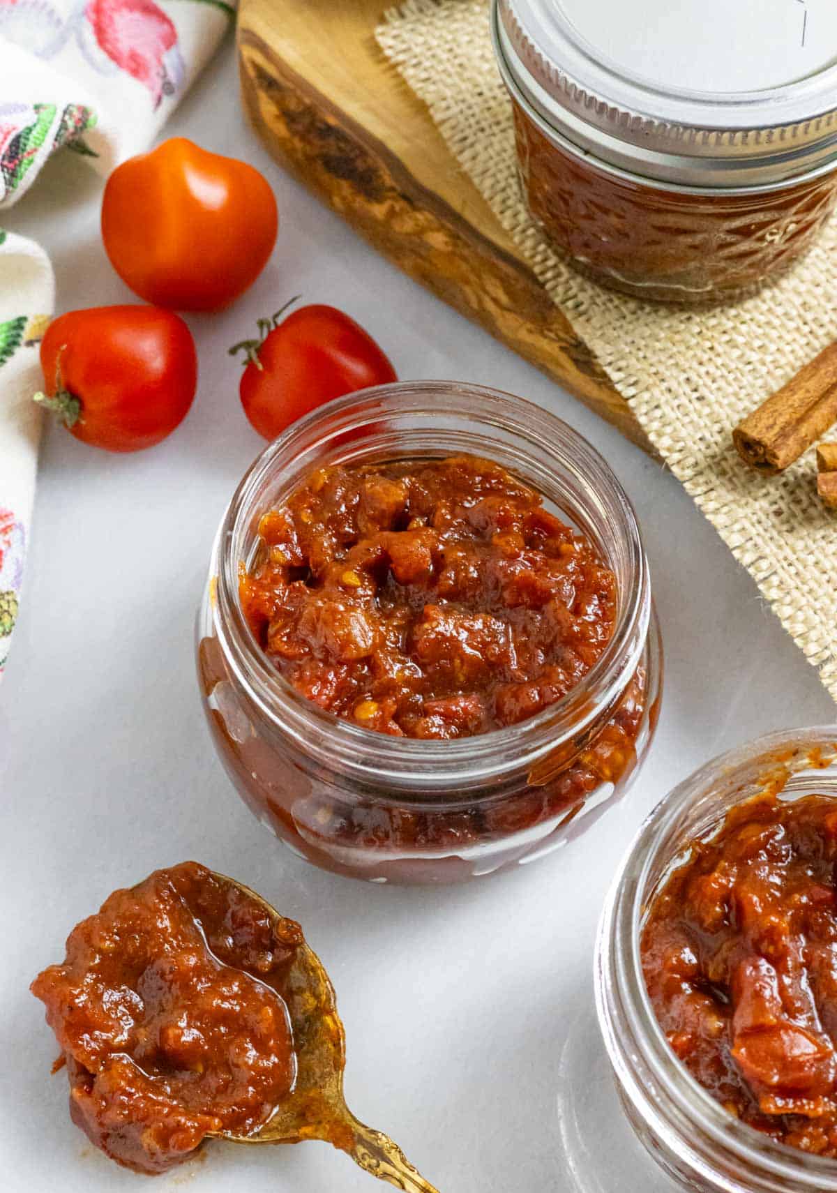 Two clear jars of tomato jam on a marble board with 3 tomatoes.