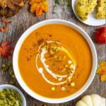 White bowl of carrot and pumpkin soup topped with spices, pumpkin seeds and a drizzle of coconut milk next to a small plate of cheddar biscuits.