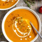 Two white bowls of carrot pumpkin soup with a spoon in one, topped with a swirl of coconut milk, pumpkin seeds and spices.