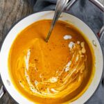 Pot of pumpkin soup with carrot with coconut milk and spices on top and a spoon in the pot.