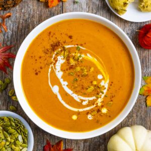 Bowl of carrot and pumpkin soup garnished with seeds, spices and a drizzle of coconut milk on a rustic board.