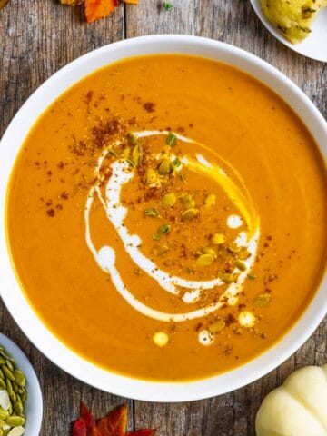 Bowl of carrot and pumpkin soup garnished with seeds, spices and a drizzle of coconut milk on a rustic board.