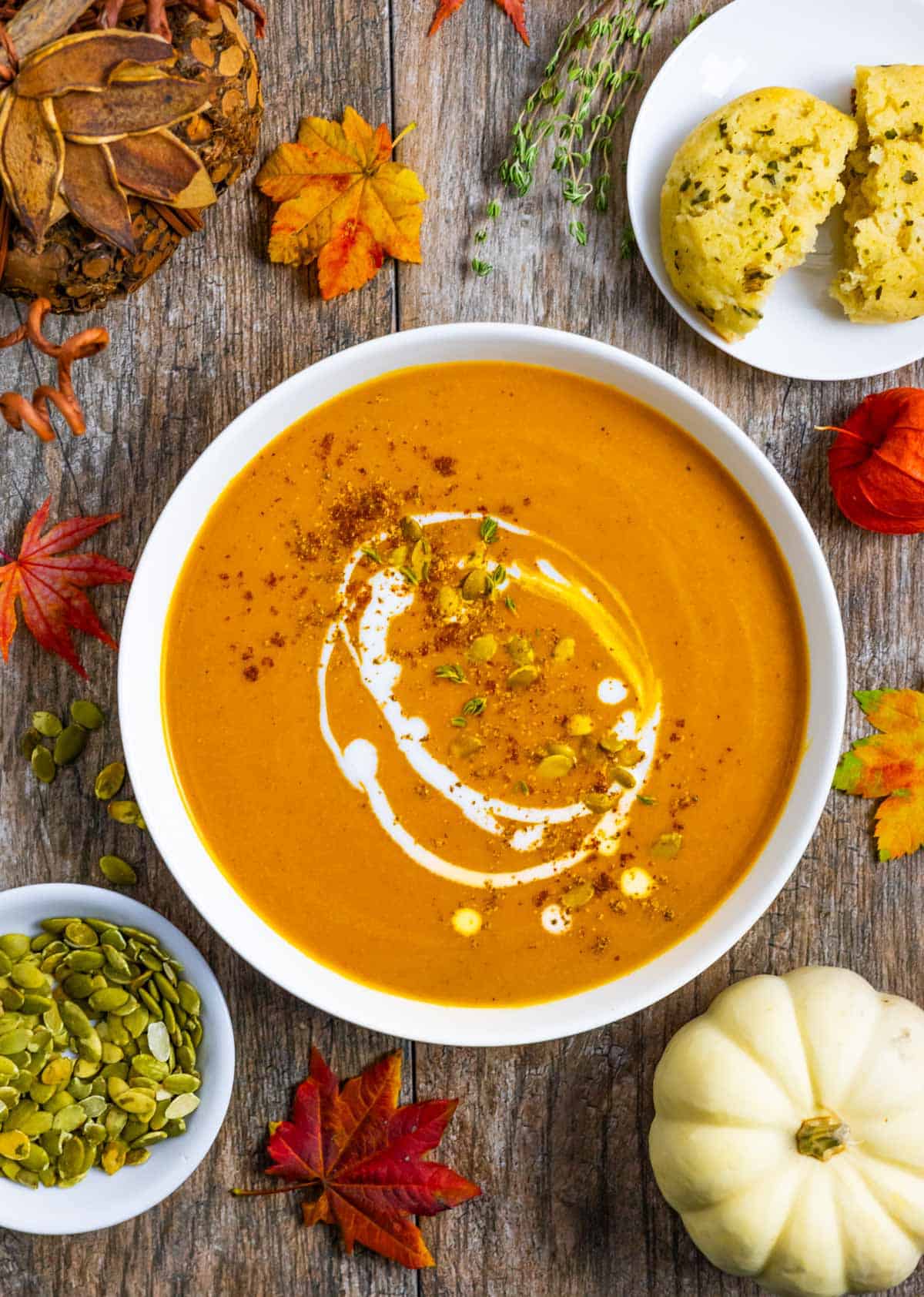 White bowl of carrot and pumpkin soup topped with spices, pumpkin seeds and a drizzle of coconut milk next to a small plate of cheddar biscuits.