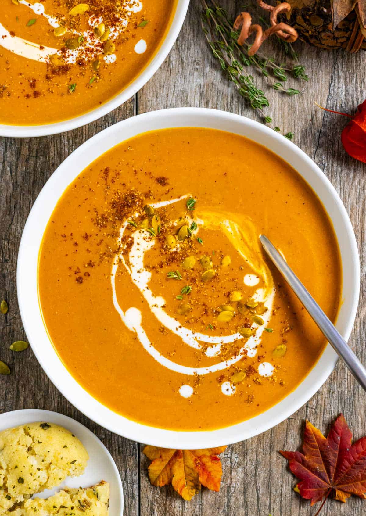 Two white bowls of carrot pumpkin soup with a spoon in one, topped with a swirl of coconut milk, pumpkin seeds and spices.