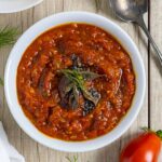 Bowl of tomato onion chutney with herbs on top on a wood board with a spoon next to the bowl.