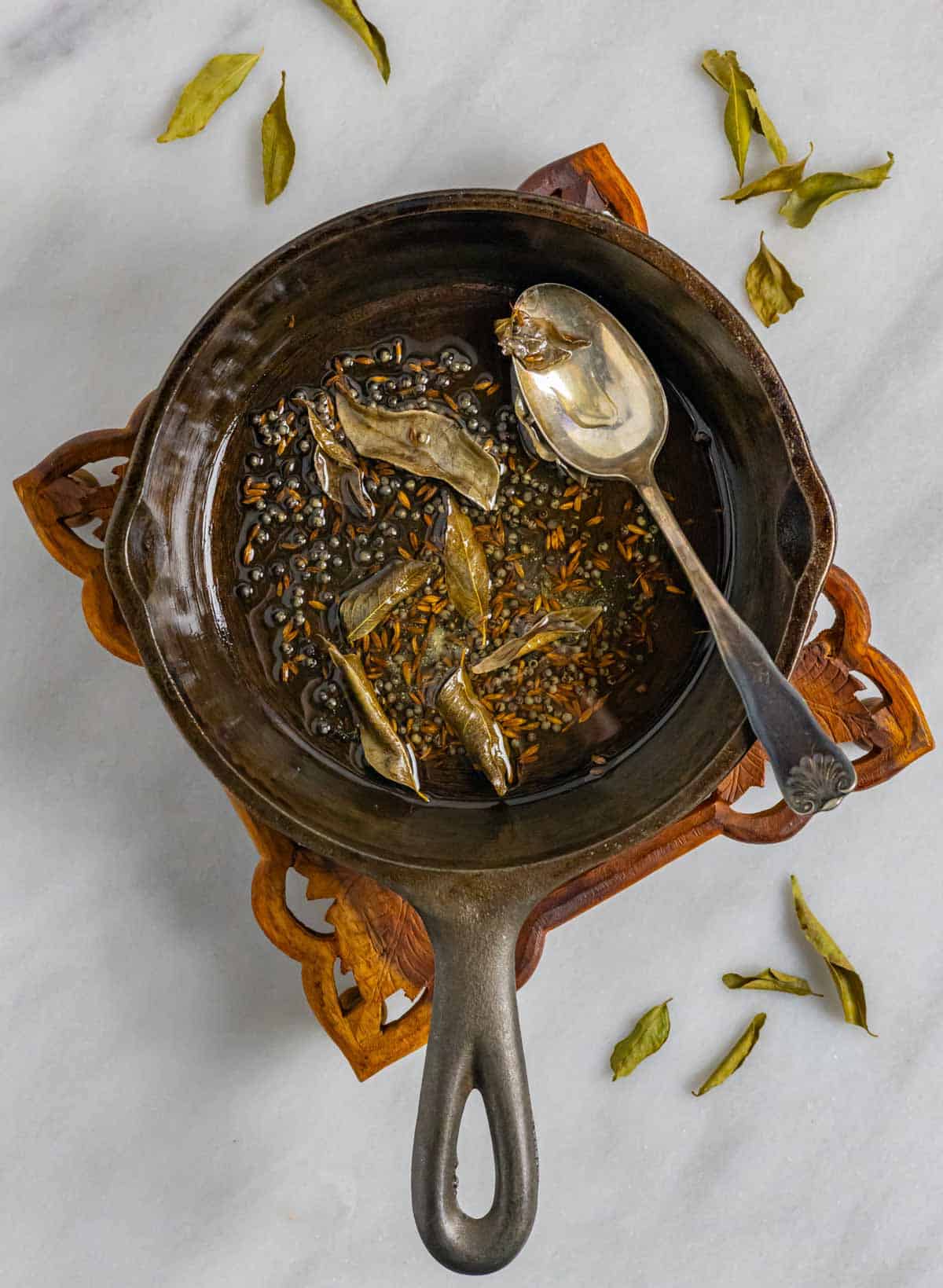 Small cast iron skillet with tempered herbs on a decorative wood trivet on a marble board.