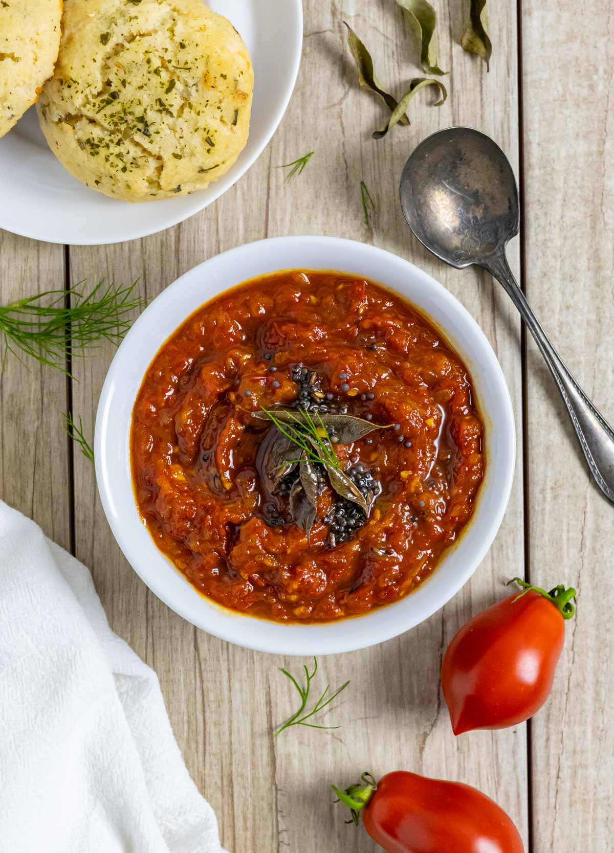 White bowl of onion tomato chutney topped with herbs on a board with a spoon, whole tomatoes, and a plate of chedar biscuits.