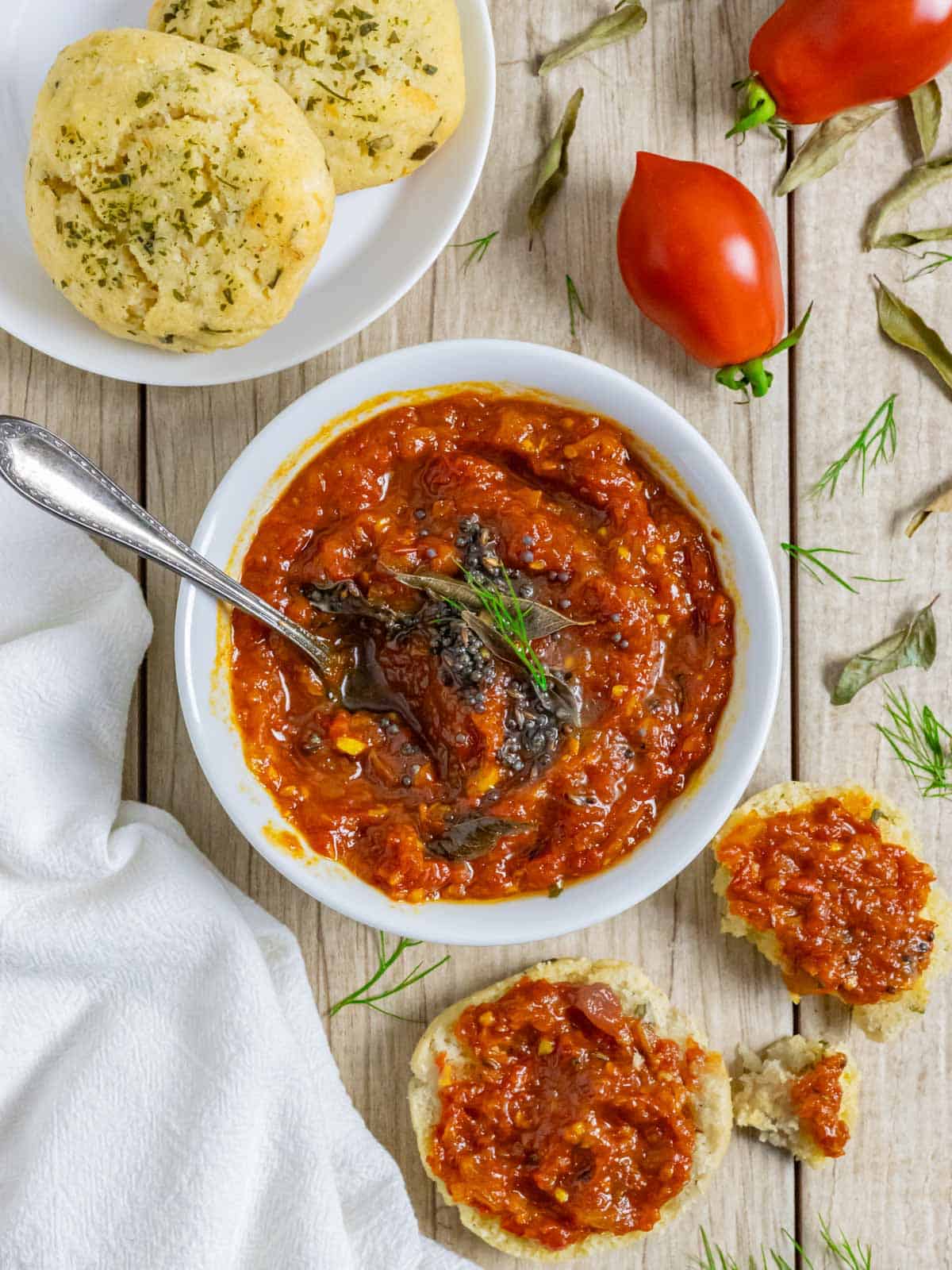 Tomato and onion chutney in a white bowl with a spoon, next to cheddar biscuits spread with chutney.