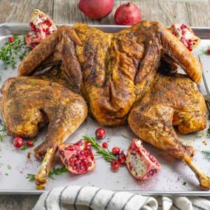 Whole spatchcock smoked turkey on a silver tray with herbs and pomegranate halves.