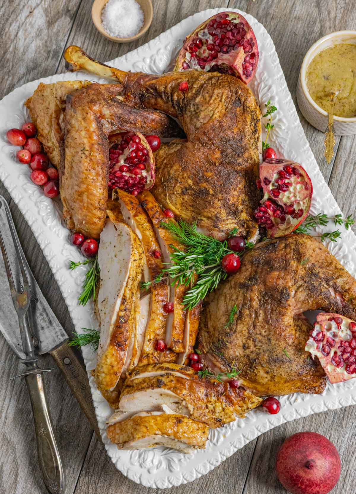 Smoked turkey breast slices, wings, leg quarters on a serving tray with herbs, cranberries and pomegranate halves.