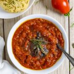 Bowl of tomato and onion chutney with a serving spoon, topped with tempered herbs on a board with whole tomatoes and a plate of chedar biscuits.