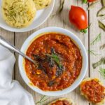 Tomato and onion chutney in a white bowl with a spoon, next to cheddar biscuits spread with chutney.