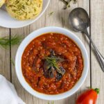 White bowl of onion tomato chutney topped with herbs on a board with a spoon, whole tomatoes, and a plate of chedar biscuits.