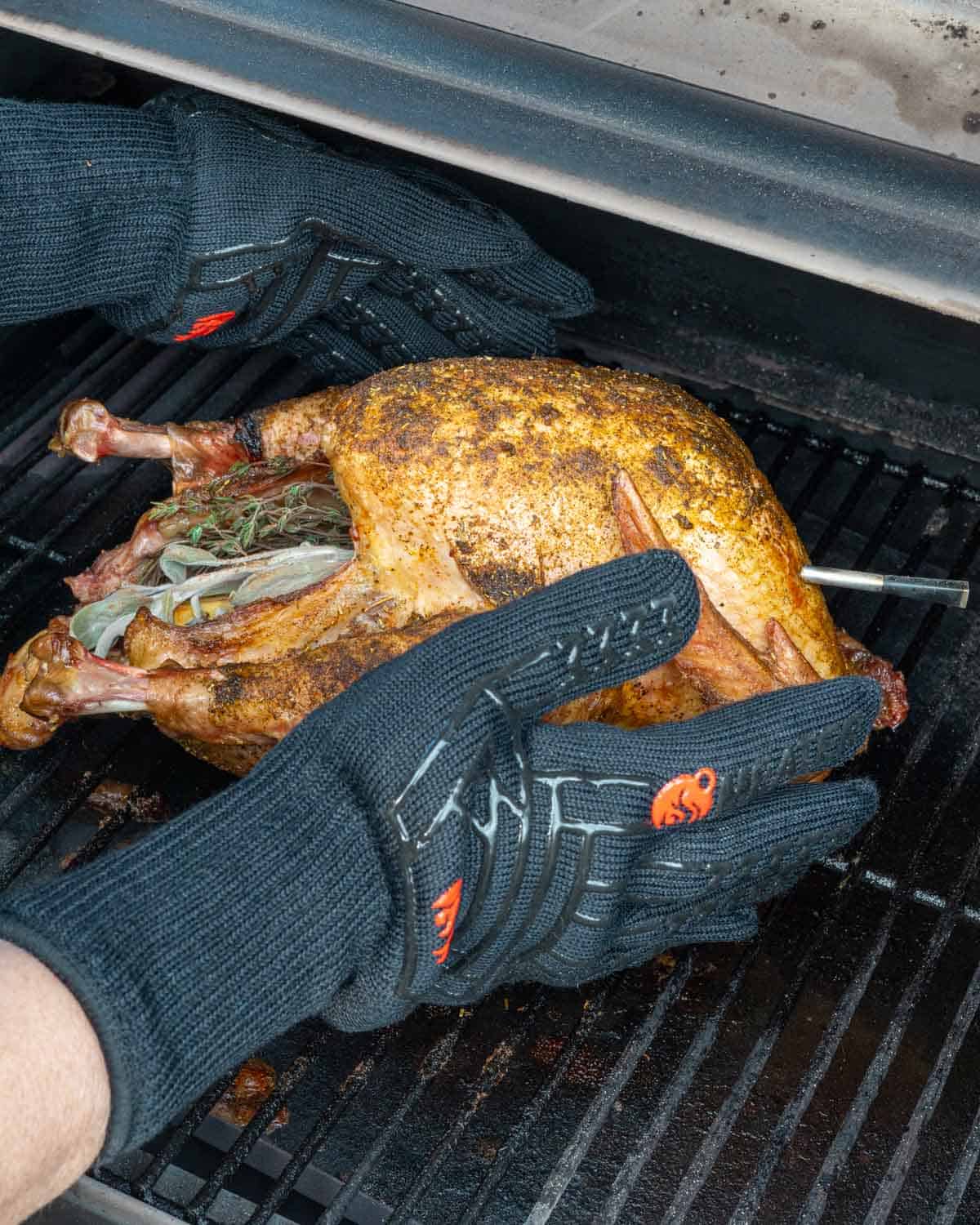 Fully smoked whole turkey being removed from smoker grates with smoker glove covered hands.