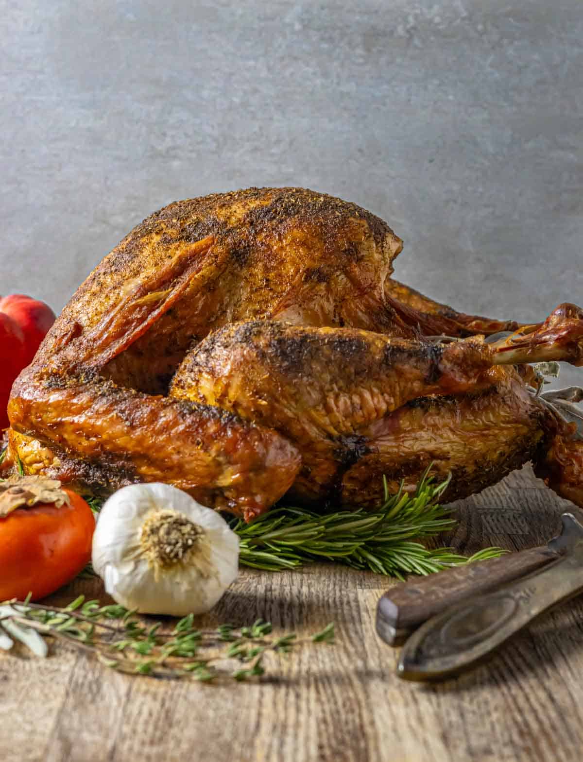 Side view of a smoked whole turkey on a board with carving knife and fork, head of garlic, herbs and a persimmon.