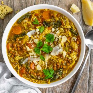 Lentil soup with turkey in a white bowl topped with shaved parmesan and parsley on a wood board with a spoon next to it.