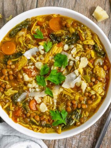 Lentil soup with turkey in a white bowl topped with shaved parmesan and parsley on a wood board with a spoon next to it.