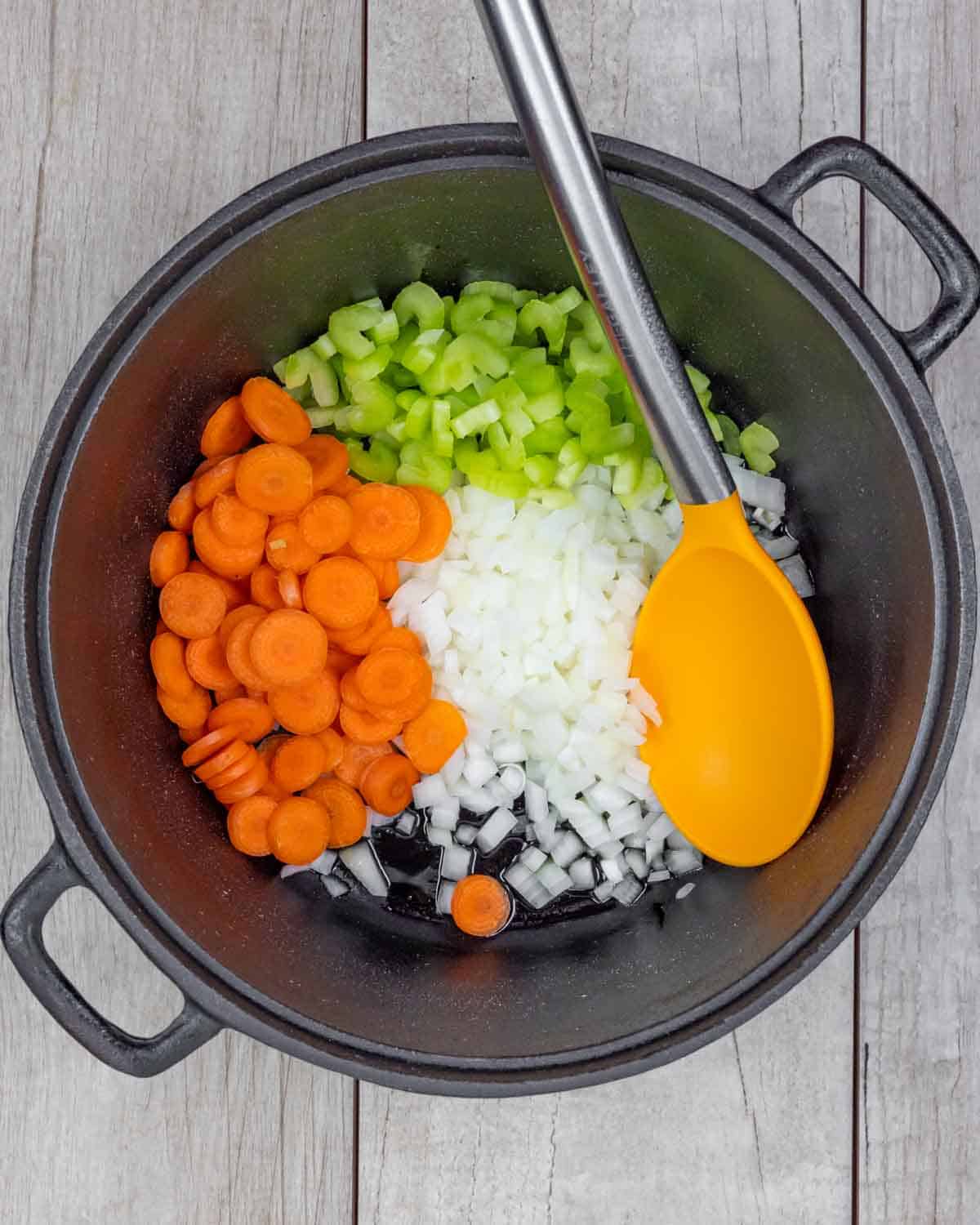 Dutch oven with raw carrots, onions and celery ready to saute with a large spoon in the pot.