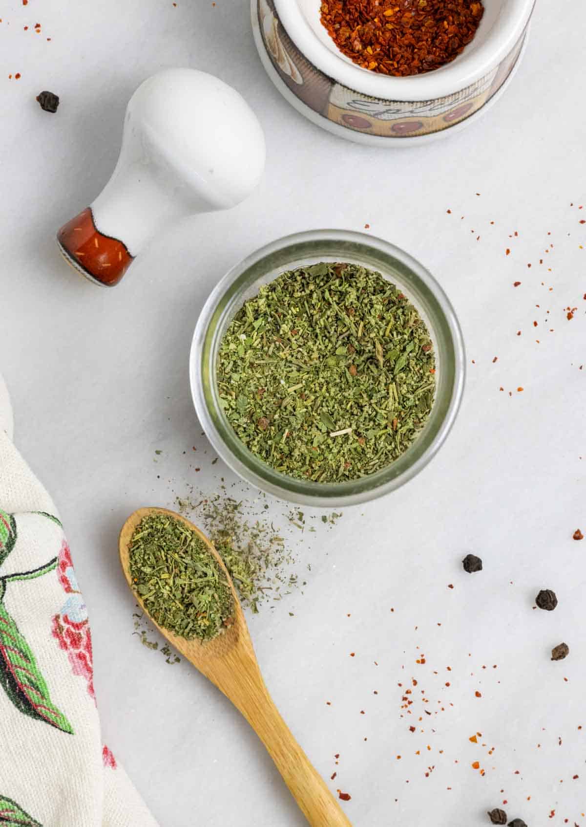 Jar of mediterranean seasoning mix on a white board with a wood spoon of spice next to it and a small mortar and pestle.