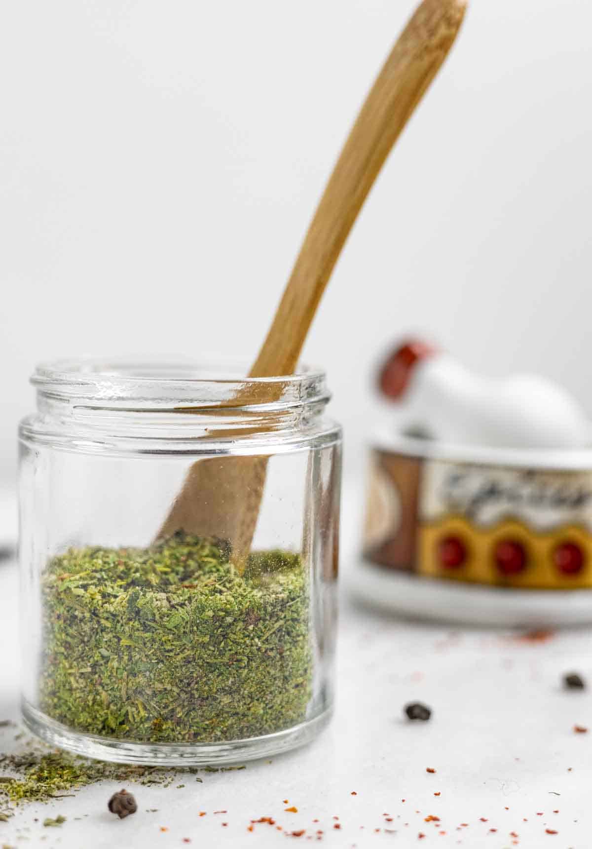 Small jar of greek spice mix with a small wood spoon in it on a marble board with spices scattered on it.