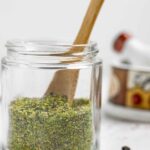 Small jar of greek spice mix with a small wood spoon in it on a marble board with spices scattered on it.