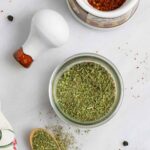 Jar of mediterranean seasoning mix on a white board with a wood spoon of spice next to it and a small mortar and pestle.