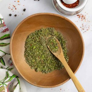 Mediterranean spice mix in a wood bowl with a small wood spoon in it on a white marble board with spices scattered on it.