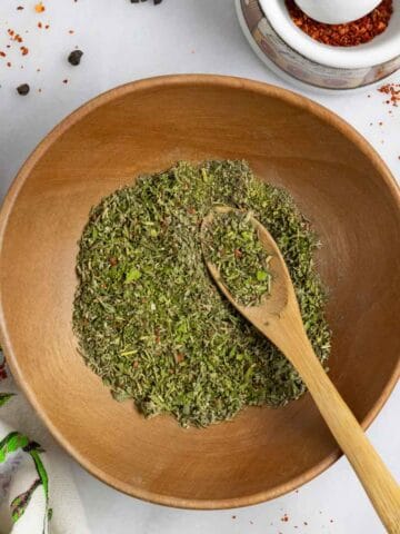 Mediterranean spice mix in a wood bowl with a small wood spoon in it on a white marble board with spices scattered on it.