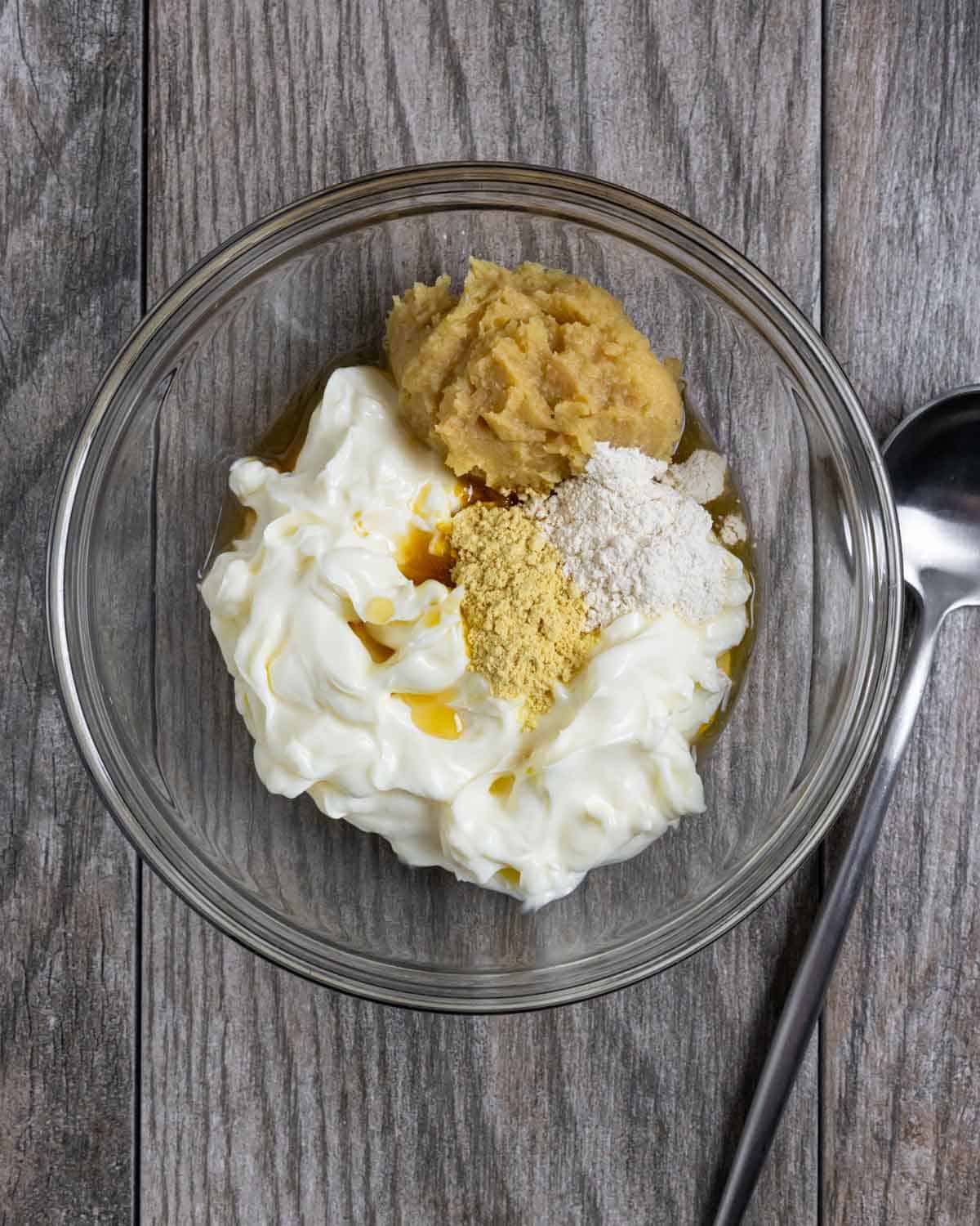 Clear glass bowl with small piles of miso mayo ingredients ready to be mixed, on a board with a spoon.