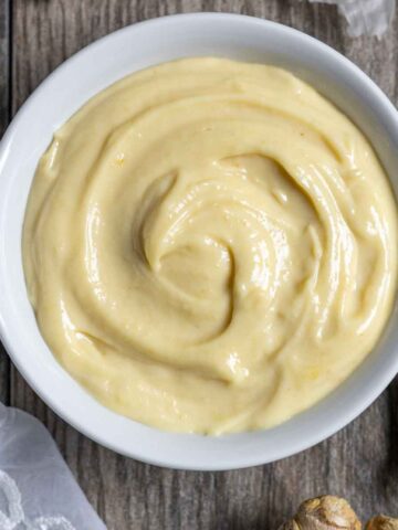 Creamy miso mayonnaise swirled in a white bowl on a board with whole garlic cloves and ginger.