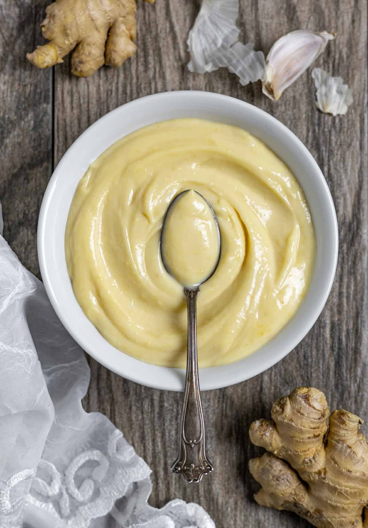 White serving bowl of miso mayo with a fancy spoon of mayo floating in the center of the bowl, on a board with whole ginger pieces.