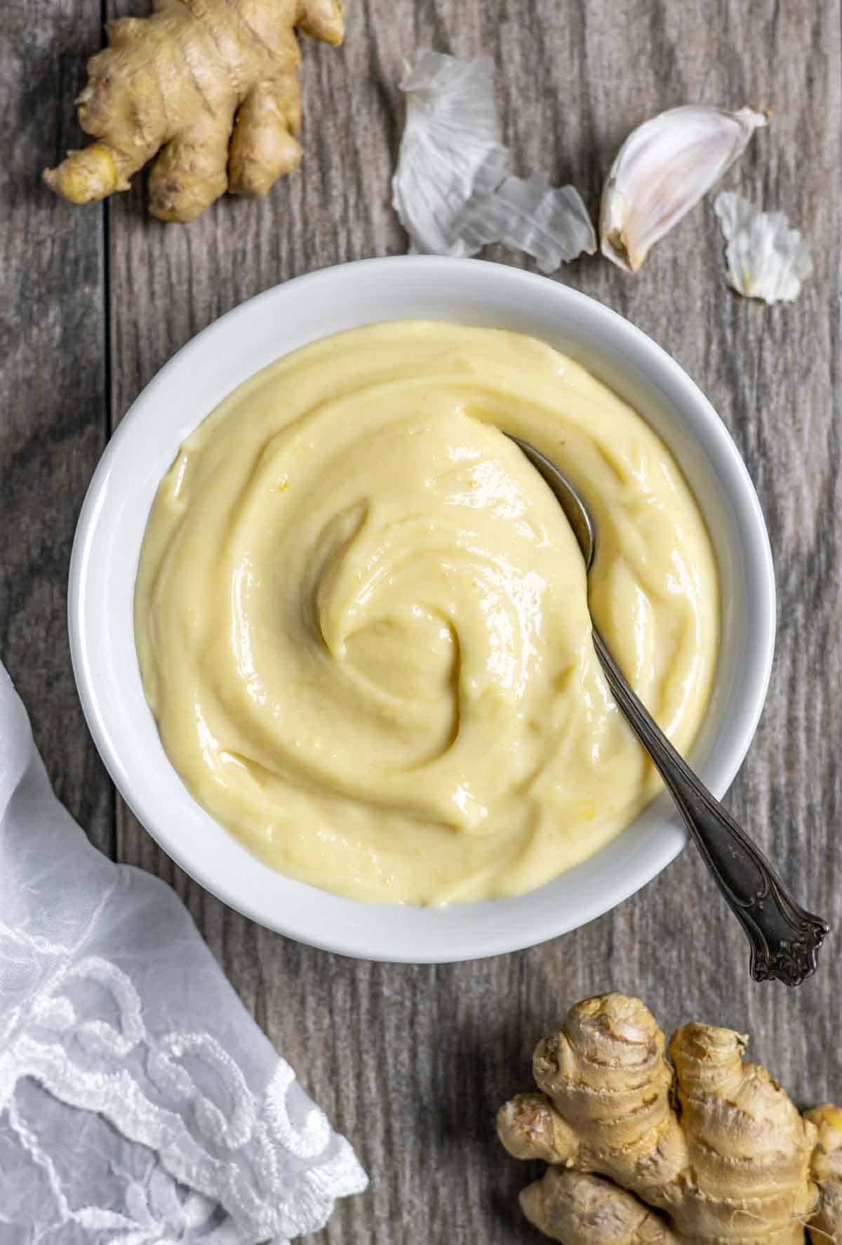 White bowl of miso mayonnaise with a spoon in it, on a board with whole ginger root, whole garlic clove and white lace.