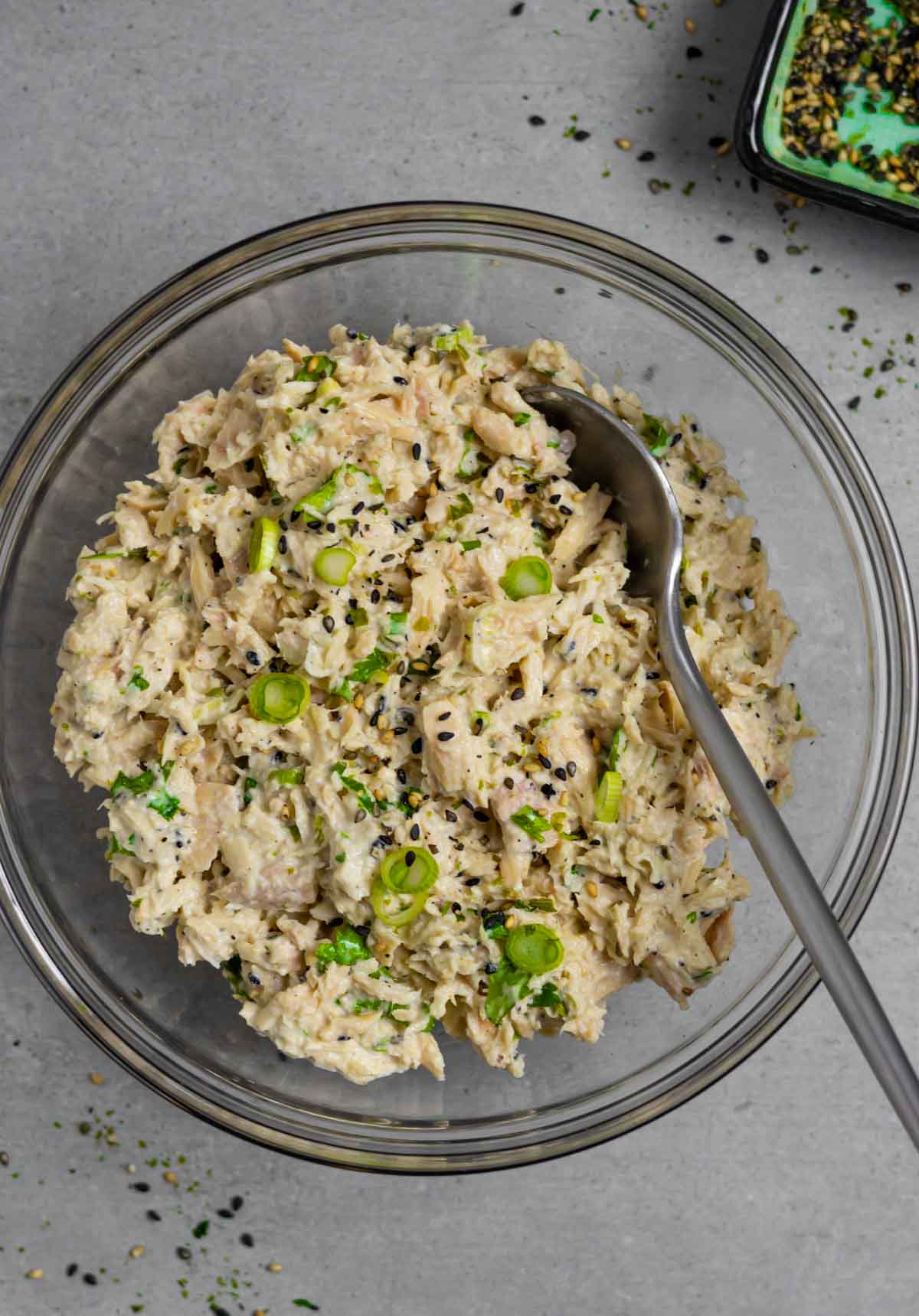 Bowl of miso tuna salad with a serving spoon in it on a grey board with a small bowl of black and white sesame seeds.