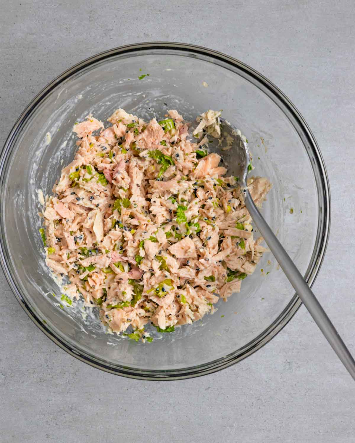 Glass bowl with mixed miso mayo tuna salad and a spoon on a grey board.