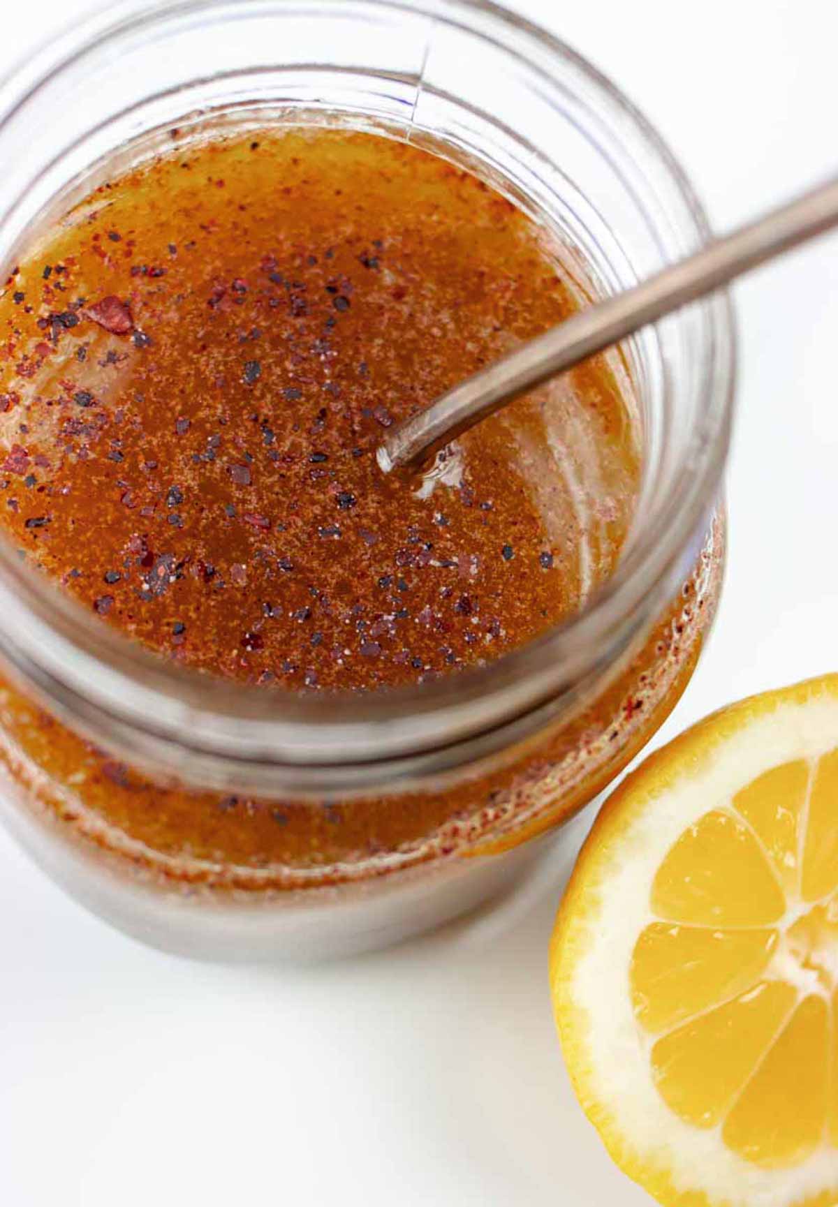 Looking into top of small mason jar of sumac dressing with a spoon in it and a lemon slice next to it.