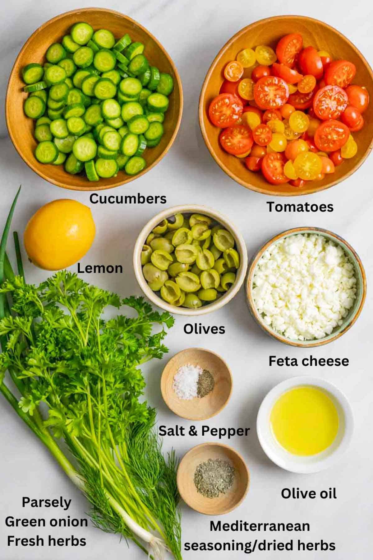 Fresh ingredients for greek salad in bowls on a board with black text labels.